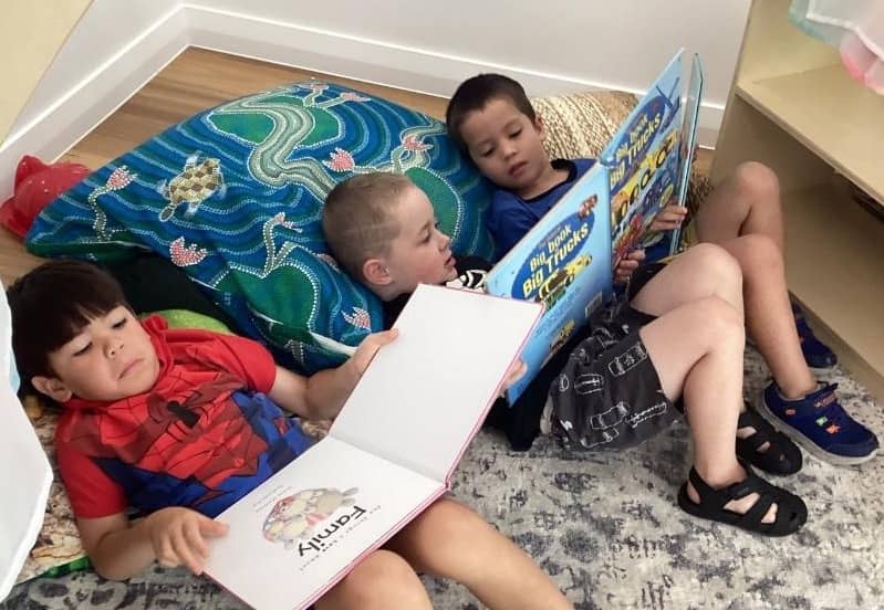 Three young children reading books together on a vibrant Aboriginal-themed cushion, sitting on a soft rug with visible hybrid flooring in the background, creating a cozy and engaging reading space.