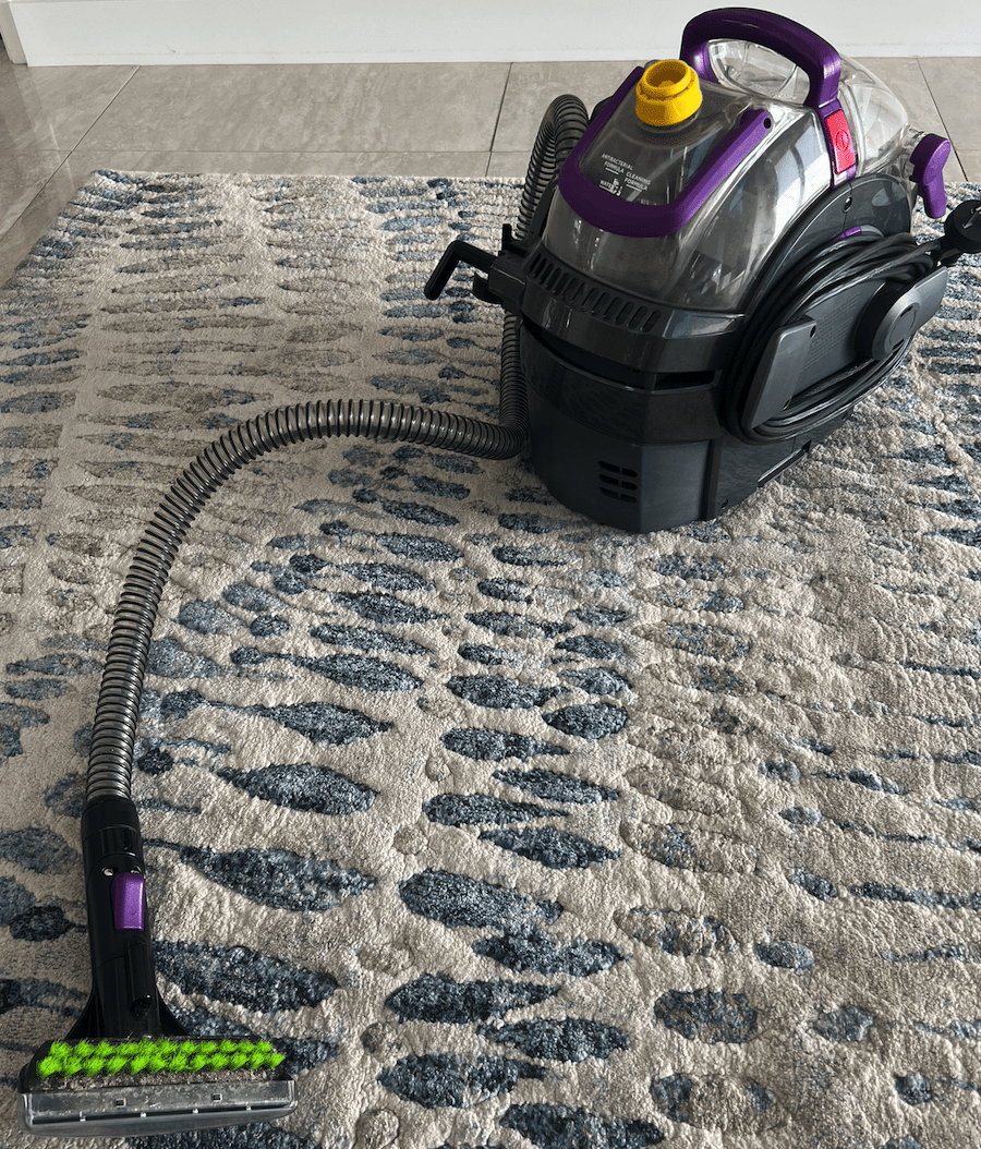 Compact Bissell carpet cleaner with purple accents and green brush attachment being used on a cream and blue patterned area rug on light tile flooring