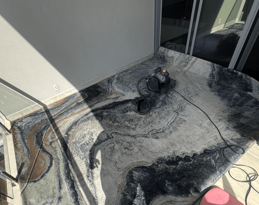 Compact Bissell spot cleaner sitting on a large marble-effect area rug in grey, black and brown tones during the machine cleaning process on an outdoor balcony - demonstrating effective deep cleaning for modern synthetic rugs