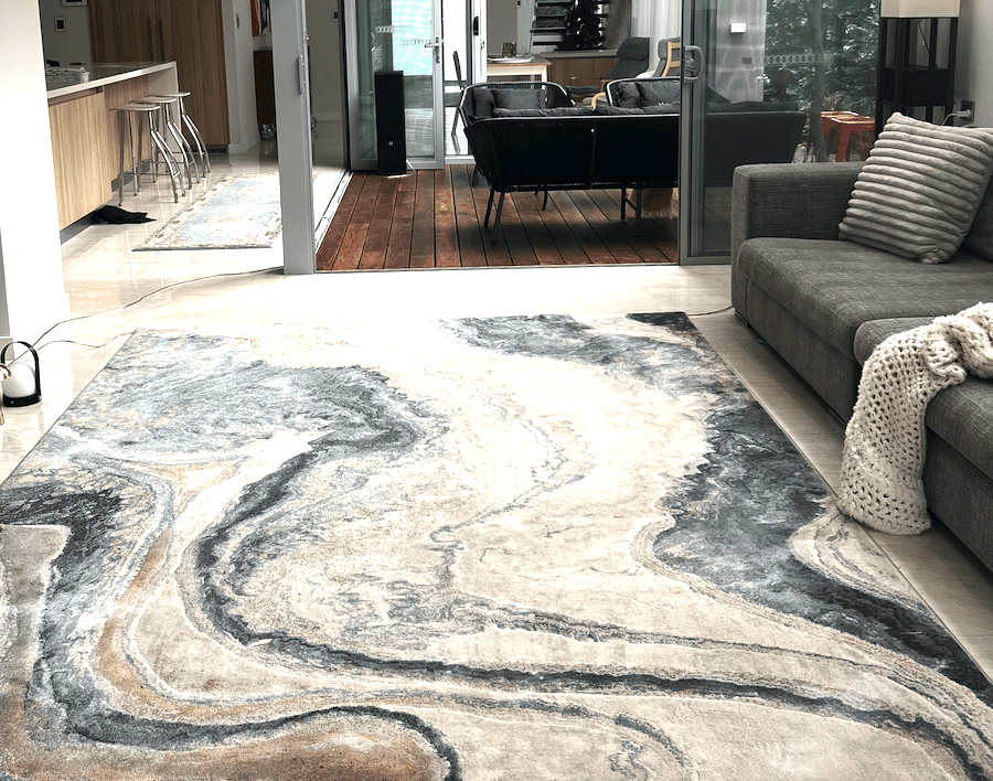 Large freshly cleaned marble-effect area rug with swirling grey, cream and beige patterns placed in a modern living room with grey sofa and wooden deck visible through glass doors