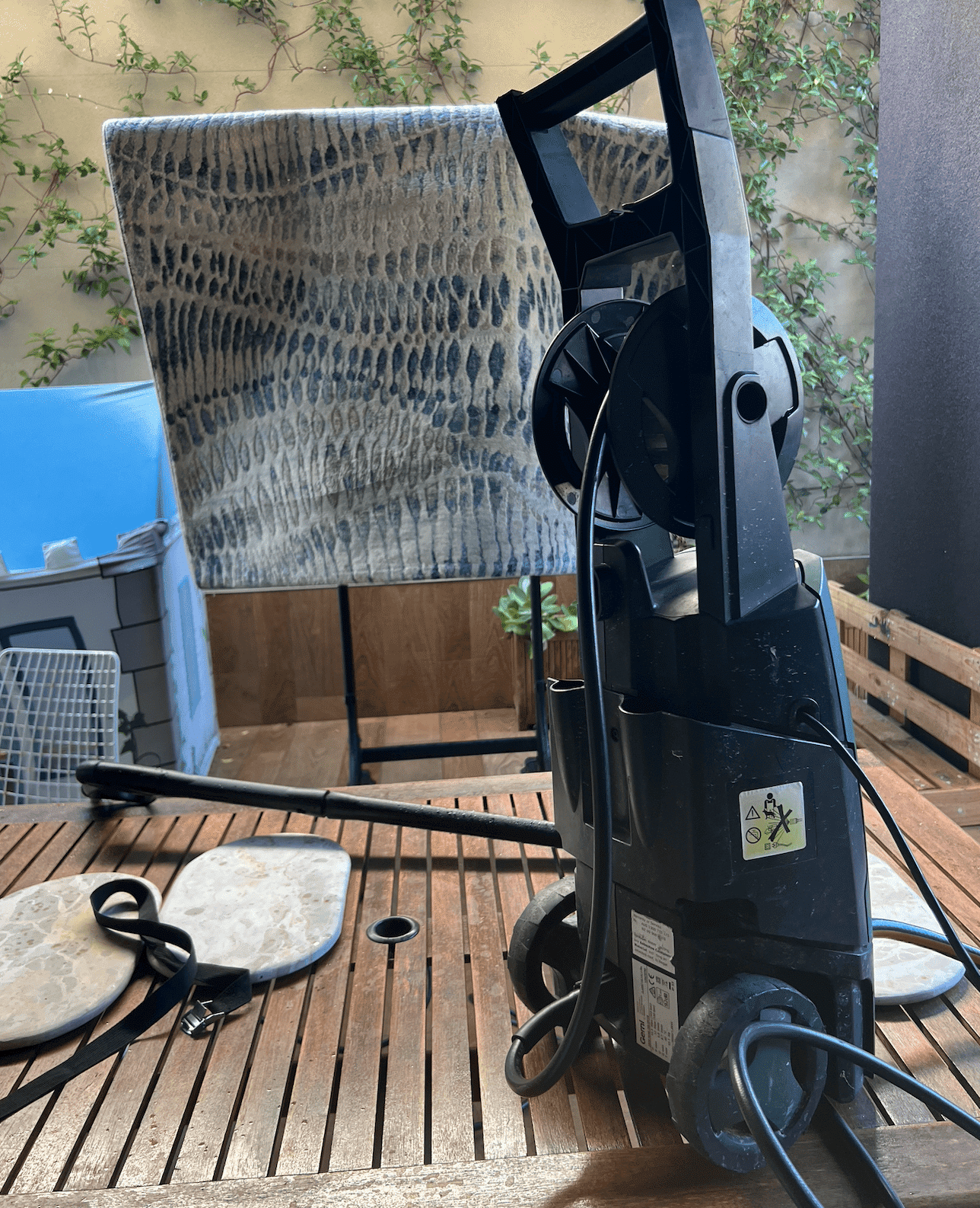 Pressure washer on a wooden deck with a blue and beige patterned rug hanging to dry in the background, demonstrating the setup for pressure washing rugs outdoors